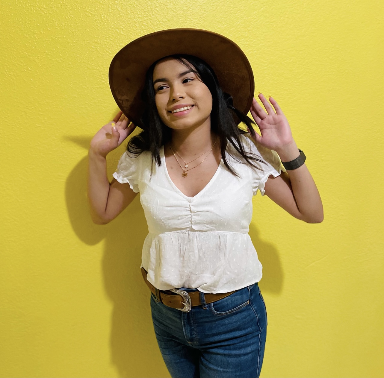 Female smiling with shoulder length black hair reaching up toward a wide brimmed hat, white blouse and jeans. The background is a bright yellow.