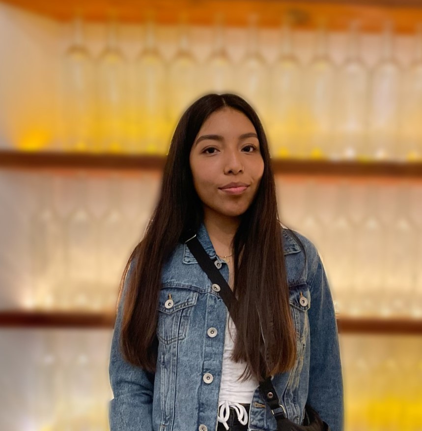 Janet is female smiling in a white shirt, jean jacket and black long strap purse. The background is blurry with two brown shelves, yellow lights and clear bottles.